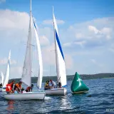 Regata żeglarska na jeziorze. Kilka łodzi z załogami ściga się, mijając zieloną bojję.