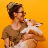Uśmiechnięta dziewczyna z okularami i bandaną na głowie tuli corgi. Piesek liże ją po nosie. Wiele imion na N, jak Nela czy Nika, pasowałoby do tej słodkiej pary.