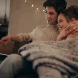 Para młodych ogląda coś na laptopie, przytuleni pod kocem. Ich bliskość i uśmiechy sugerują, że historia o "fake dating" tak bardzo angażuje emocjonalnie czytelników, bo pozwala przeżywać rodzące się uczucia.