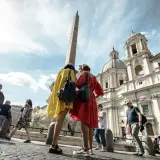 Turisti ammirano l'obelisco e la chiesa a Roma ad agosto, con il sole che illumina la piazza.