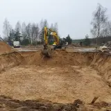 Żółta koparka pracuje przy wykopy pod fundamenty. W tle drzewa i biały van.