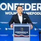 Mężczyzna w garniturze przemawia na tle logo Konfederacji. Czy to zapowiedź sekretów negocjacji Konfederacji z rolnikami w sprawie blokad granic w marcu 2026?