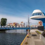 Niebieski most na łebskiej promenadzie, z widokiem na żaglowiec i charakterystyczną, futurystyczną budowlę.