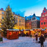 En snötäckt vinterviken julmarknad med ett stort, upplyst träd och färgglada hus. Folk strosar bland bodarna.