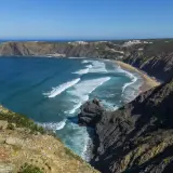 Malownicze wybrzeże Portugalii z klifami, piaszczystą plażą i turkusowym oceanem. Białe fale rozbijają się o brzeg, a w oddali widać białe domki.