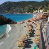 Plage animée à Moneglia, Italie, avec des parasols rayés, des baigneurs dans la mer turquoise et des collines verdoyantes en arrière-plan.