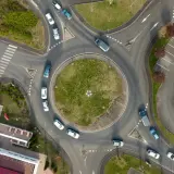 Widok z lotu ptaka na rondo, które jest skrzyżowaniem z ruchem okrężnym. Samochody poruszają się po okręgu, a obok znajduje się parking.
