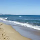 La playa de Xeraco, con arena dorada y el mar azul, se extiende hacia un horizonte con edificios y montañas.