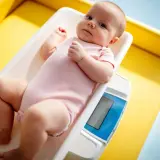 Noworodek w różowym body leży na wadze. Rodzice często zastanawiają się, ile powinien przybierać noworodek, by mieć pewność, że rozwija się prawidłowo.