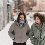 Deux amies discutent en marchant dans la neige. Une idée outfit hiver parfaite : doudoune beige et manteau vert ceinturé.