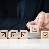 Dłoń kładzie kostkę z krzyżykiem na tle innych z zaznaczonymi ptaszkami. To symbol decyzji, jak w umowie przyrzeczonej.