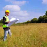 Inżynier w kasku i kamizelce odblaskowej analizuje plany na tle łąki, przygotowując się do badań geotechnicznych gruntu.