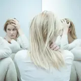 Kobieta z blond włosami w białej bluzce, trzymająca się za głowę, otoczona swoimi odbiciami w lustrze.