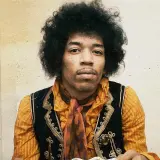Jimi Hendrix w pomarańczowej koszuli i kamizelce, z charakterystyczną fryzurą. W tle subtelna, lekko zatarte tło, jakby w **purple haze**.