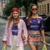 Dwie dziewczyny prezentują modny styl ubioru młodzieży: jedna w różowej kurtce i kwiecistej spódnicy, druga w sportowej koszulce i wzorzystej spódniczce.