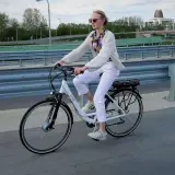 Kobieta w białych spodniach i sweterku jedzie na rowerze elektrycznym. W tle most i budynki.