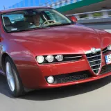 Czerwone Alfa Romeo 159 1.9 JTD pędzi po drodze, kierowca skupiony na trasie.