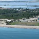 Vista aérea de la costa de El Saler, con el mar azul, la playa, dunas, vegetación, edificios y salinas al fondo.