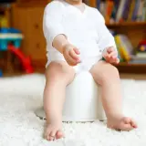 Niemowlę na nocniku, być może z powodu biegunki. W tle zabawki i książki.