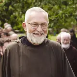 Bruder Paulus, ein älterer Mann mit Brille und Bart, lächelt in die Kamera. Hinter ihm stehen weitere Männer in braunen Kutten.