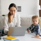 Kobieta pracuje na laptopie, podczas gdy jej syn rysuje obok. To codzienna opieka na dziecko i praca zdalna w jednym.
