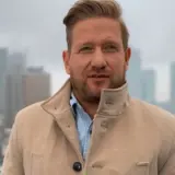 Bartek Jędrzejak w płaszczu na tle miasta. Marzy o powrocie do żony i dzieci po pracy.