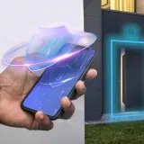 Dłoń trzyma smartfon z ikoną tarczy, symbolizującą bezpieczeństwo. Obok drzwi domu świeci się niebieska ramka, sugerując, że keyless entry to nowoczesny sposób na wejście.