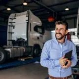 Uśmiechnięty mężczyzna z tabletem w ręku, w tle warsztat z ciężarówkami. Oferuje **usługę transportową ryczałt**.