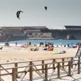 Plaża pełna kolorowych latawców i ludzi, gotowych do **kitesurfingu w Polsce**. W tle widać zabytkowe budowle.