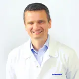 Uśmiechnięty mężczyzna w białym fartuchu lekarskim na jasnym tle.