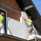Dwóch budowlańców montuje płyty styropianowe, wykonując termoizolację budynku.