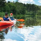 Dwie osoby płyną kajakiem po spokojnej rzece, otoczonej zielenią. Idealne miejsce na spływ kajakowy Mazury, gdzie najlepiej poczuć bliskość natury.