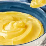 Una ciotola blu piena di deliziosa crema pasticcera vegana, con un cucchiaio che ne preleva una porzione.