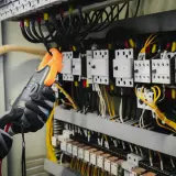 Ręce w rękawicach trzymają pomarańczowy miernik, badając skomplikowany opis rozdzielni elektrycznej z licznymi przewodami i przekaźnikami.