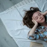 Junge Frau mit geschlossenen Augen und Händen an den Schläfen auf einem Bett liegend. Ein Buch liegt daneben. Tipps für gesunden Schlaf.