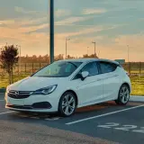 Biała Opel Astra K zaparkowana na parkingu. Słońce zachodzi, tworząc ciepłą atmosferę. Czas na opel astra k wymiana akumulatora.