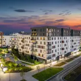 Nowoczesne osiedle w Warszawie Ursus o zmierzchu. Jasne budynki mieszkalne z balkonami i oświetlonymi oknami, otoczone zielenią i alejkami.