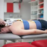 Pacjent leży na kozetce, poddawany zabiegowi z użyciem prądów Traberta. Elektrody przymocowane są do jego pleców.