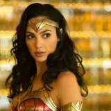 Gal Gadot jako Wonder Woman, część obsady filmu, w złotej zbroi i opasce na głowie, na tle rozmytych, złotych świateł.