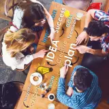 Zespół przy stole dyskutuje nad planem, rozwijając swoją kreatywność. Słowa "IDEA", "plan", "success" i "goal" tworzą mapę myśli.