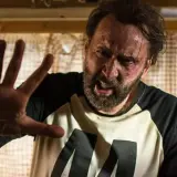 Nicolas Cage med blåmärken och svettig panna, höjer handen i en gest av stopp. En scen från en av hans filmer.