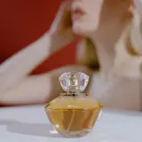 Elegancka buteleczka perfum z płynem o barwie **citral** na białym tle, w tle niewyraźna sylwetka kobiety.