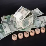 Dom z banknotów 100 zł, symbolizujący kredyt hipoteczny. Słowo "KREDYT" ułożone z drewnianych pieczątek.