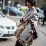 Kobieta w szarym żakiecie trzyma torbę z monogramem Gucci, jakby to był certyfikat autentyczności.