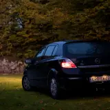 Czarny Opel Astra H 1.6 benzyna zaparkowany na trawie, w tle jesienne drzewa.