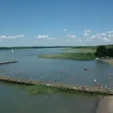 Wypływa z jeziora roś, a plażowicze kąpią się i opalają. W oddali widać żaglówki i łodzie.