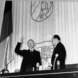 Ein Mann hebt die Hand, neben ihm ein weiterer Mann. Im Hintergrund die deutsche Fahne und Wappen. Der ältere Bundeskanzler spricht.