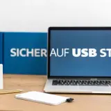 Laptop mit "Sicher auf USB Stick" auf dem Bildschirm. Ein weißer USB-Stick und eine externe Festplatte liegen auf dem Schreibtisch.