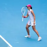 Iga Świątek w akcji, w białej sukience i różowej czapce, gotowa do uderzenia. Jej buty sportowe idealnie pasują do stroju.