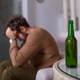 Mężczyzna z głową w dłoniach siedzi obok pustej butelki piwa. Może to symbolizować uczulenie na alkohol lub jego nadużywanie.